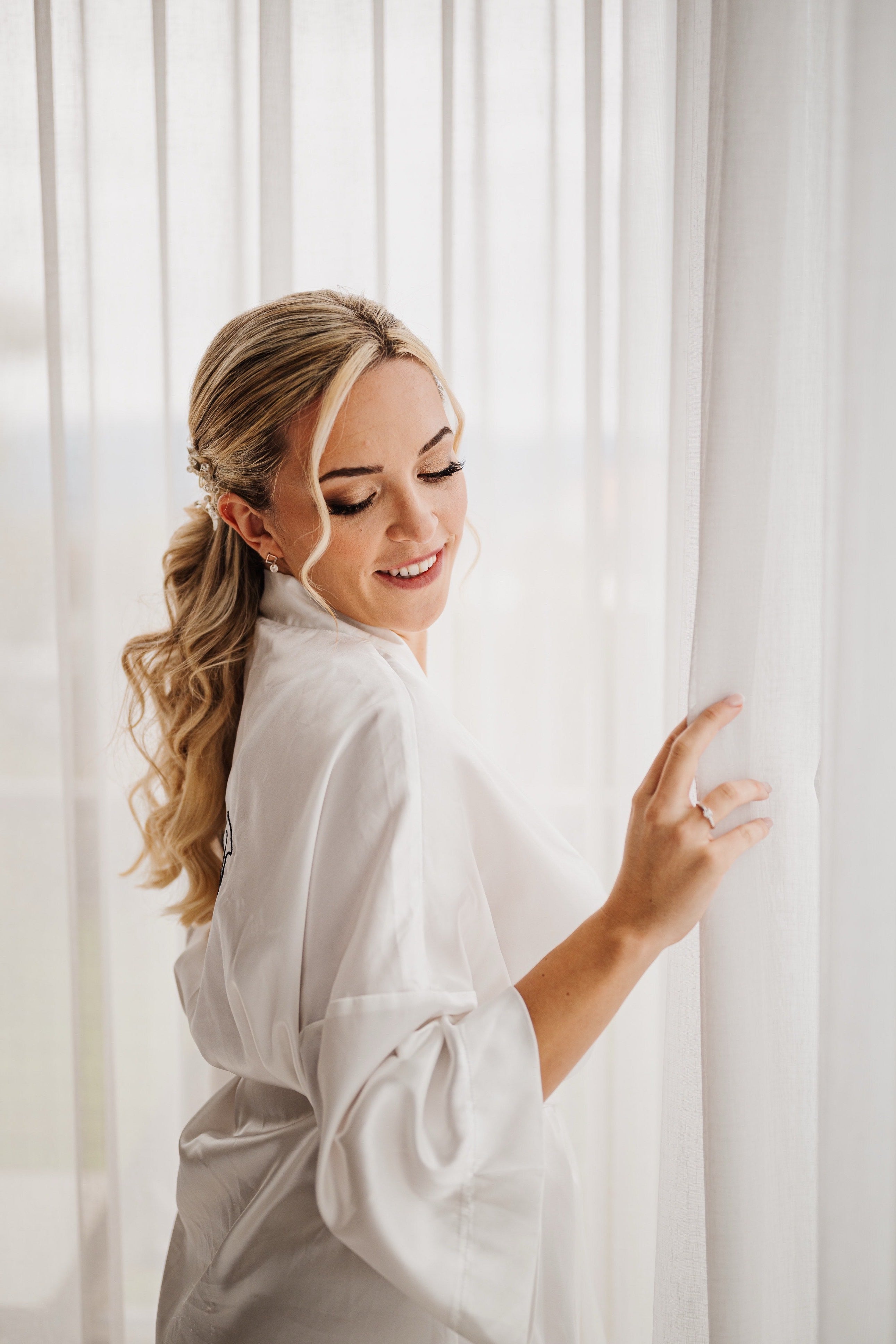 A woman with blonde hair in a half-up ponytail wearing a white satin robe, smiling and looking over her shoulder while standing in front of white sheer curtains.